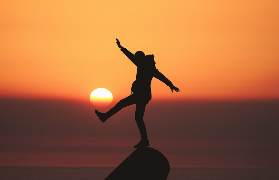 A silhouette of a woman jumping on top of a rock at sunset.